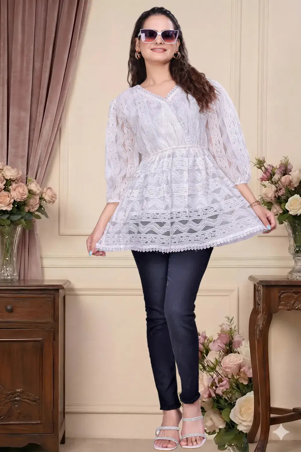 Woman wearing a white lace top and black pants in a decorated room with flowers and furniture.