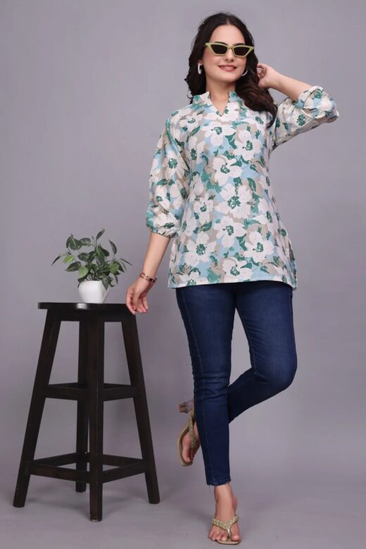 Model wearing a teal and white floral tunic with blue jeans, posing beside a black stool.